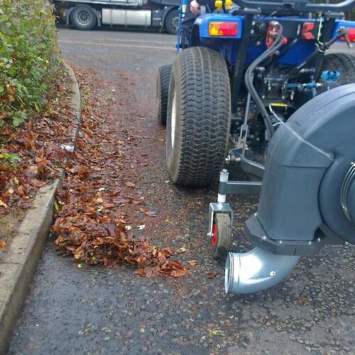 Tractor-mounted blower clearing leaves along a kerb